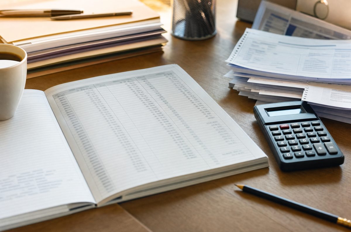Libro de contabilidad abierto con columnas de cifras, junto a una calculadora, un lápiz y reportes impresos.